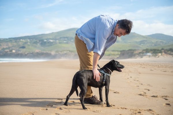 Ein Mann steht mit seinem schwarzen Hund an der Leine am Strand und legt ihm die Hand auf die Flanken